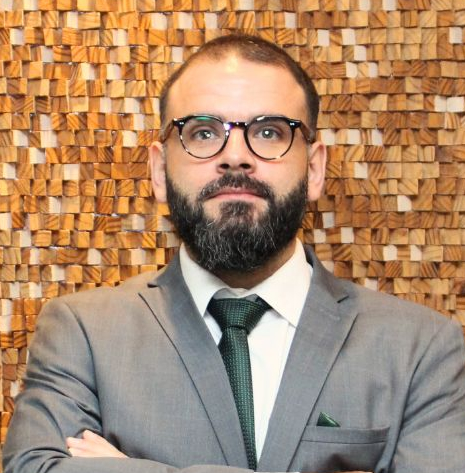 Marcello Brandão - Homem de óculos e barba olhando para a frente com os braços cruzados