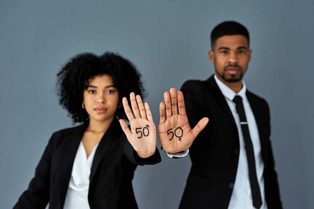 Imagem de um home e uma mulher, ambos negros, vestindo terno preto e camisa branca. Eles estão com as palmas da mão para frente e nelas há um número "50" com o símbolo de masculino e feminino.