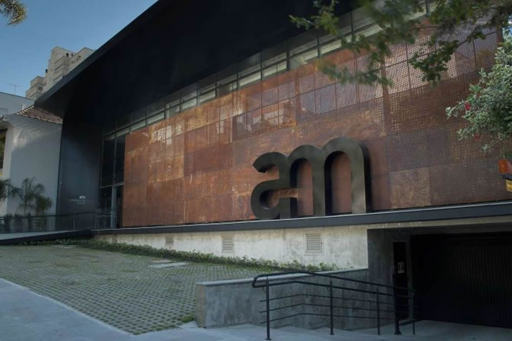 A imagem apresenta um detalhe da fachada de um edifício moderno. O destaque
fica por conta de um grande logotipo, composto pelas letras "AM", em um estilo
minimalista e com contornos arredondados. As letras, confeccionadas em metal
preto, contrastam com o fundo oxidado e com textura, que reveste toda a parede.
A iluminação indireta, que realça as letras, cria um efeito visual marcante e
confere à fachada um aspecto sofisticado e contemporâneo. A composição da
imagem sugere um ambiente urbano e cosmopolita, com um design que valoriza a simplicidade e a elegância.